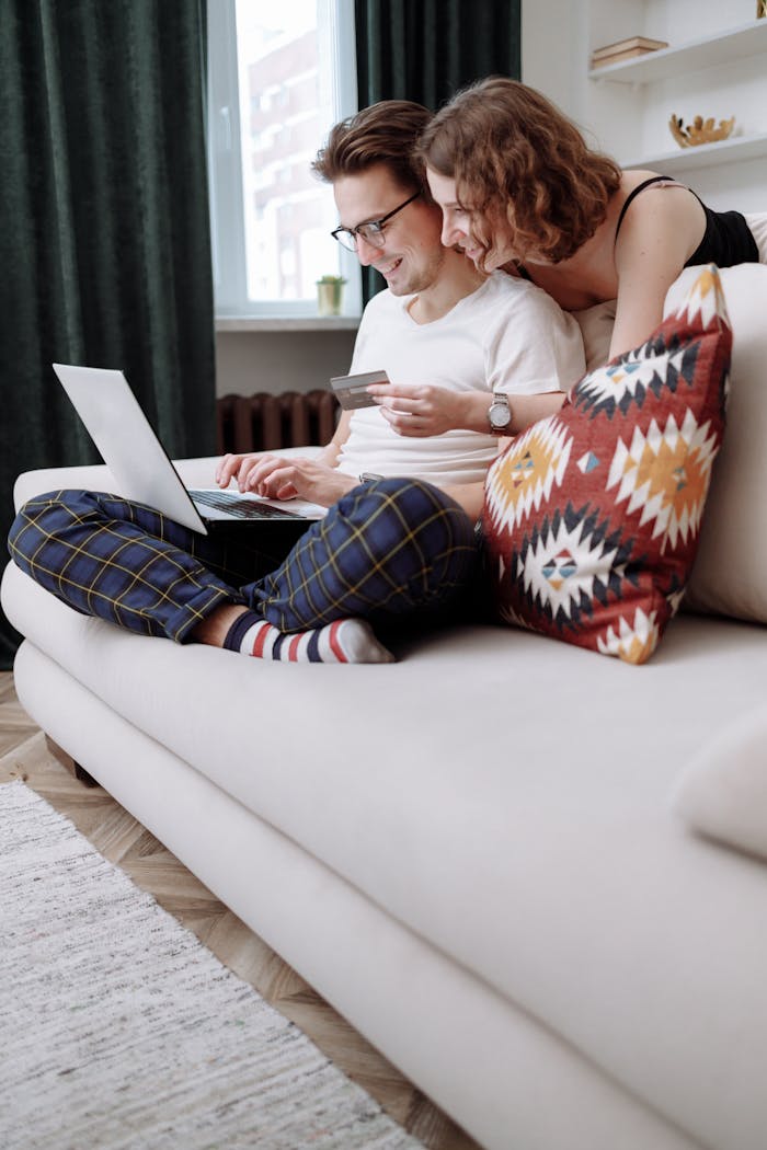 services-01 Happy couple sitting on a couch, using a laptop for online shopping with a credit card in hand.